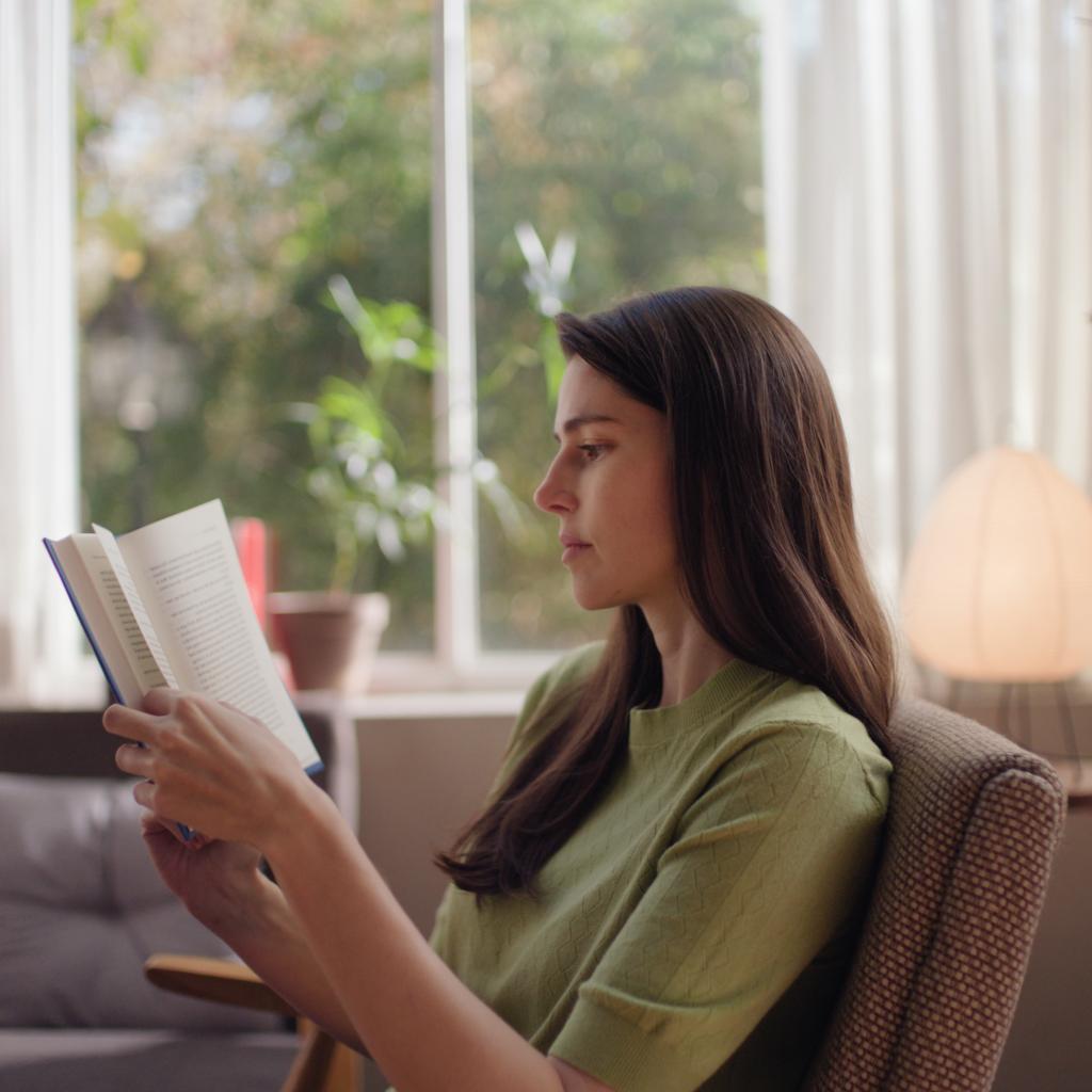 Osoba czyta książka w przytulnym fotelu przy oknie z kubkiem herbaty obok